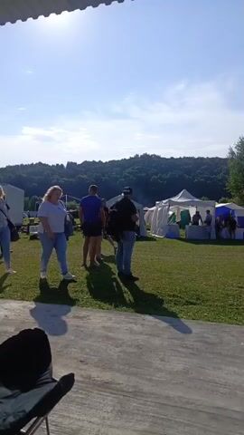 The video shows an outdoor event with several tents and people gathered in a grassy field under a clear blue sky with some clouds. In the foreground, there is a concrete area with a portable grill. Several people are visible in the mid-ground, some standing and talking, others walking. In the background, there are more tents and a large crowd of people. The overall scene suggests a festival, fair, or outdoor market. The lighting indicates it is daytime.