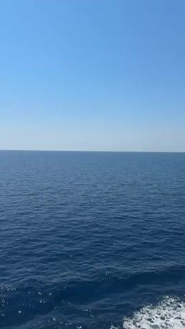 The video shows a view from a boat moving through the sea. The water is a deep blue, and there are waves. The sky is clear and blue. In the distance, there is a landmass with hills. The boat's railing and a portion of its superstructure are visible on the right side of the frame. White foam trails behind the boat, indicating movement.