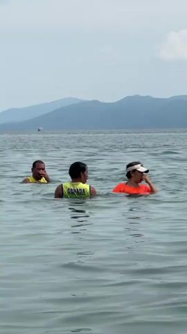Five individuals are waist-deep in calm ocean water under a partly cloudy sky. In the foreground, a person wearing a yellow and green "GERADA" vest talks with a person in an orange shirt and white visor. To their left, another person in a yellow shirt watches. To their right, a person in a pink bucket hat and another person in a dark shirt are present. The individuals are interacting and appear to be smiling and laughing. Distant mountains or hills are visible on the horizon, with a small boat or object in the far background.