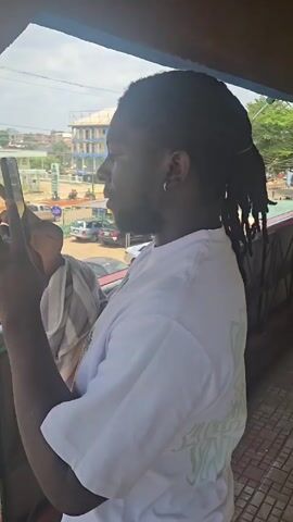 A man with dark skin and shoulder-length dreadlocks pulled back in a ponytail stands on a balcony, looking out at a street scene. He wears a white t-shirt with a colorful graphic design and a silver hoop earring in his left ear. He holds a black phone in his left hand and makes a gesture with his right hand, appearing to be talking. A white towel rests on his left arm. Behind him on the balcony, a man in a red shirt walks by. The street below is visible, showing cars, buildings, and people. The overall setting is bright and appears to be outdoors during the day.
