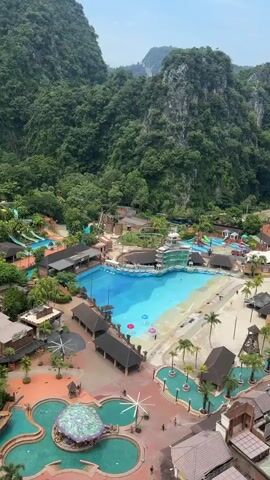This is an aerial drone shot of a water park surrounded by lush green mountains. The water park features several pools, including a large wave pool with a sandy beach area. There are also water slides, lounge chairs, and palm trees visible. The scene is busy with people enjoying the water park. The weather appears to be sunny.