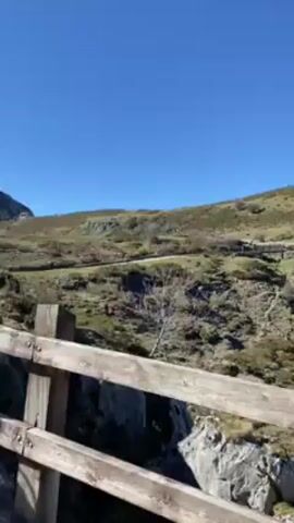 The video shows a panoramic view of a rocky, mountainous landscape under a clear blue sky. The foreground features a wooden fence, suggesting the perspective is from a viewing point. The landscape is rocky with small vegetation, and the sky is clear. The camera slowly pans across the landscape, revealing more of the rocky terrain and vegetation.