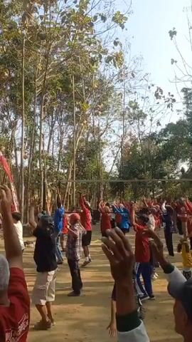 A group of people, varying in age, are outdoors participating in what appears to be a coordinated exercise routine. They are arranged in rows on a dirt or sand surface, with trees and foliage in the background. Many of the individuals are wearing red or white clothing. They are performing arm movements, with most of their arms raised upwards. A volleyball net is visible behind the group. There are also flags visible in the background, including what appears to be an Indonesian flag. The sky is clear and bright.