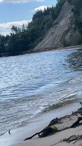 The video shows a calm lake with gentle waves lapping against a sandy shore. A steep, vegetated cliff rises in the background, with a trail or staircase visible on its side. The sky is partly cloudy, with the sun casting a soft light on the scene. The water is a deep blue, and the sand is a light brown. There are some fallen branches and debris on the shore in the foreground. The overall impression is one of natural beauty and tranquility.