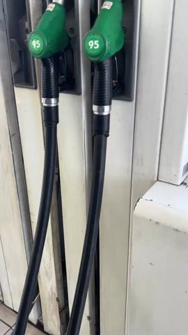 The video shows a person at a gas station, using a gas pump to fill up their car. The gas pump is white and has a green handle labeled "95." A pair of blue disposable gloves hang next to the pump. Keys sit on top of the pump as well. The display on the gas pump shows an increasing amount of money, suggesting that gas is being dispensed. The car being fueled is a dark-colored SUV with a spare tire mounted on the back. The surrounding environment includes a paved gas station lot with other vehicles and buildings visible in the background.