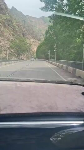 A car travels along a road, a view from the dashboard. The road is surrounded by mountains and trees. A fence runs alongside the road on the right. A few other cars are visible in the distance. The sky appears overcast.