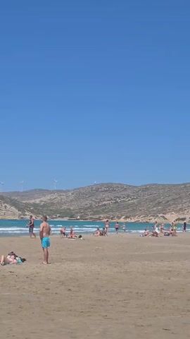 A wide shot of a busy beach on a sunny day. The sand is light brown and there are several people scattered across it, some lying down, some standing, and some walking. The ocean is a bright turquoise color with waves breaking on the shore. There are several kite surfers in the water, their colorful kites visible against the clear blue sky. In the background, there are hills covered in scrubby vegetation and a few wind turbines. The sky is a vast expanse of clear blue.