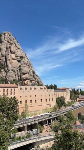The video shows a scenic view of a monastery complex situated on a mountainside, against a clear blue sky with scattered white clouds. The monastery buildings are made of light-colored stone, with architectural details like arched windows and tiled roofs. A road or viaduct is visible, with cars and people moving along it. The mountain, with its distinctive rock formations, is partially covered in greenery. Vegetation is also present in the foreground, framing the scene. The sky is bright, suggesting it is daytime.