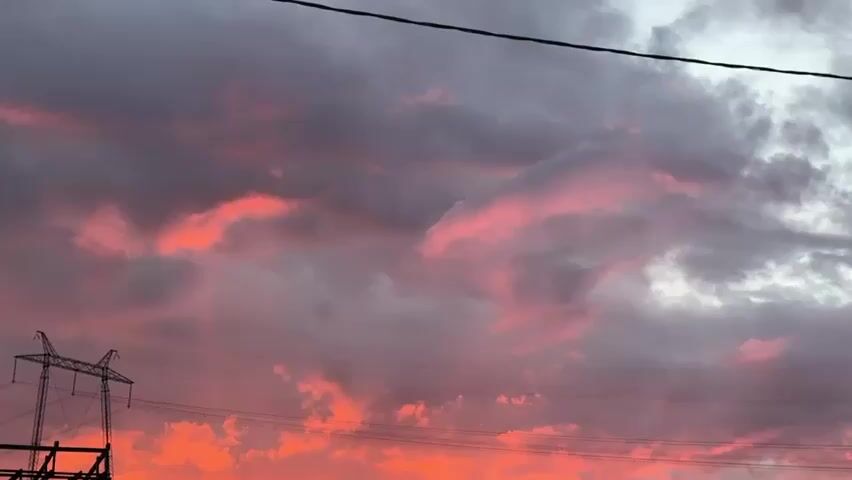 This is a short video of a young girl with long, dark hair, wearing a gray hooded sweatshirt. She is looking off to the side with a pensive expression. The background shows a grassy field and a dark fence. In the upper portion of the video, the sky is filled with dramatic clouds illuminated by a vibrant pink and orange sunset. There are also power lines and a power pylon visible against the colorful sky. The camera angle is somewhat low, looking up towards the sky and the girl.