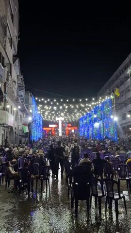 The video shows an outdoor night event with a large crowd of people seated and standing. A stage is set up at the far end of the street, adorned with bright lights and what appears to be a large, illuminated structure. String lights are hung overhead, illuminating the scene. The ground appears wet, suggesting recent rain. People are casually dressed and many are facing the stage. The atmosphere seems festive and crowded, with security personnel visible among the attendees.