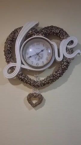The video shows a close-up, slightly angled shot of a decorative wall clock. The clock is shaped like a heart with the word "LOVE" integrated into its design. The word "LOVE" is written in large, stylized, white lettering, with the "L" and "O" forming part of the heart's outline, and the "V" and "E" positioned inside the heart. The heart itself is bordered by a textured, ornate pattern, appearing to be made of small, dark gold or bronze-colored rosettes. In the center of the heart is the clock face, which is round and white with black Arabic numerals. The clock has two hands, a minute hand and an hour hand, both black. A small, dangling heart ornament, matching the color and style of the border, hangs from the bottom center of the clock via a thin chain and a decorative piece. The background is a plain, textured wall, light beige or cream in color. The lighting is even, and the focus is sharp on the clock. There are no people or any visible actions occurring in the video.