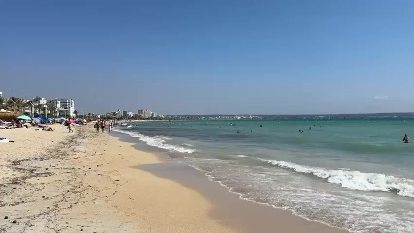 A wide shot of a crowded beach on a sunny day. The sand in the foreground is scattered with seaweed and debris. Gentle waves roll onto the shore. In the shallow water, several people are swimming and wading. Further out, more people are visible in the turquoise water. Along the coastline, buildings and palm trees line the horizon. The sky is clear and blue. Sailboats are visible in the distance.