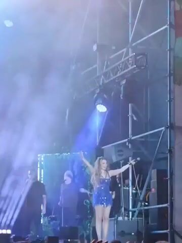 A woman in a sparkly blue dress sings and dances on a stage while a man in a black shirt sings next to her. Behind them are decorations of fake plants, equipment, and speakers. Blue lights and spotlights are shining on the stage, and the crowd is recording the performance on their phones and tablets. The person recording this video has their tablet up in the air, capturing the performance.