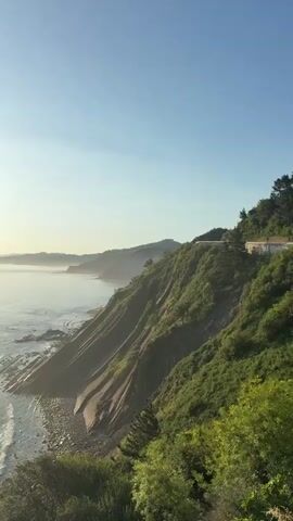 A drone shot pans across a cliffside overlooking the ocean. The sun shines brightly, casting a shimmering reflection on the water. The cliff is covered in lush green vegetation, and the ocean waves crash against the rocky shore below. The scene is peaceful and serene, with a sense of natural beauty and tranquility.