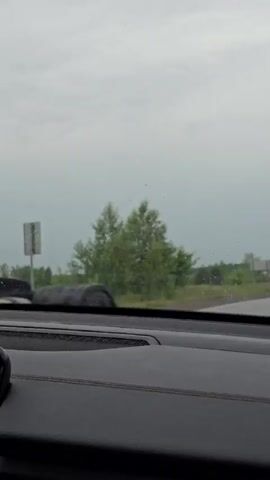 The video captures a view from inside a vehicle, looking out through the windshield. The dashboard is visible in the foreground, with stitching detail on the dark material. The weather appears overcast, with a gray sky. The scene outside consists of a grassy roadside with several trees. A dark-colored SUV with its headlights on is driving on the road ahead. Further down the road, a blue truck with a white cargo box is approaching. Traffic signs are visible along the side of the road. The camera is mounted in a way that suggests the vehicle is in motion.