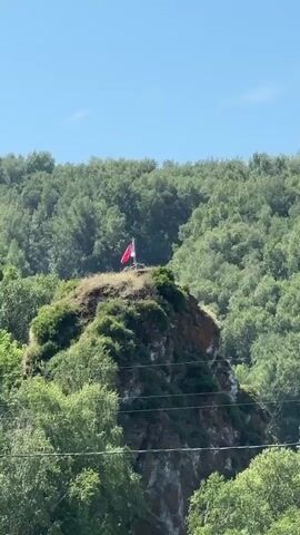 This media file captures a distant view of a rocky, tree-covered hill topped with a flag. The flag is predominantly red with a white section, attached to a flagpole. The hill is situated in a lush, green valley, with a dense forest of trees extending up the surrounding slopes and forming the background. The sky above is clear and blue, suggesting a sunny day. In the foreground, power lines are visible, hinting at human infrastructure within the natural landscape. The overall scene is peaceful and serene, evoking a sense of remoteness and natural beauty. There is no visible text within the media file.