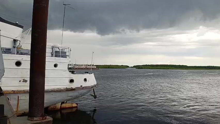 The video opens with a shot of a white boat, possibly a tugboat or small cargo vessel, docked at a pier on the left. The boat appears somewhat weathered, with rust marks visible on its hull. Its side features several porthole-like windows. The camera then pans right, revealing a wide expanse of dark, rippling water. In the distance, a narrow strip of green land, likely forested, stretches across the horizon.

The sky is heavily overcast with dark, ominous clouds, suggesting an impending storm or current inclement weather. As the camera continues to pan right, a visible curtain of rain can be seen falling over the distant land and water on the far right side of the frame. On the right bank, several industrial structures, including what appear to be cranes, are visible, indicating a port or industrial area. The overall atmosphere is somber due to the dark clouds and gray water. The scene is set in Igrim, Russia, at a waterfront location, likely a river or large lake. No visible text is present in the video.