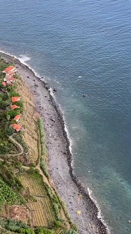 An aerial drone shot follows a coastline with a steep cliff face on the left and the ocean on the right. Terraced fields and a few houses with red roofs are visible on the green, grassy slopes. A narrow, rocky beach lines the shore, with a small pier extending into the water. The ocean is a deep blue, with some white waves breaking near the shore. The cliff face is rocky and layered, with some sparse vegetation. The drone moves slowly, panning across the scene.