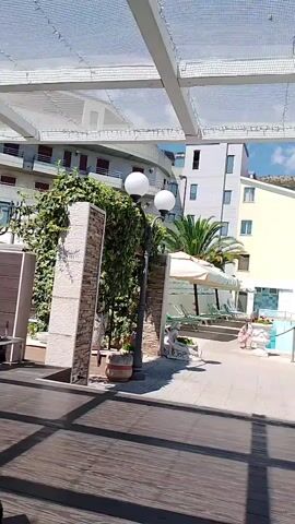 The video shows an outdoor swimming pool area of a hotel. There is a large, rectangular pool with clear blue water and lounge chairs around it. Several buildings, likely part of the hotel, are visible in the background. A palm tree is also present near the pool. The sky is blue with some clouds, indicating a sunny day. The camera pans across the scene, offering a wide view of the amenities.
