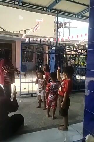 A group of five young children, dressed in casual clothing, stand in a line outdoors. They are positioned in front of a dark metal gate, which is adorned with red and white triangular flags strung across the top. The children appear to be participating in an activity, with some holding objects up to their mouths and looking upwards. One child is wearing a red shirt and brown shorts, another has on a red and white patterned shirt and shorts, and a third is in a white t-shirt and shorts. The ground in front of them is paved. In the foreground, a person is holding up a phone, seemingly recording the event. The background shows buildings and vegetation. The overall scene suggests an outdoor event or celebration.