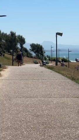 The video pans from left to right, showing a paved walkway in the foreground with grass and trees on the left. A woman is walking on the walkway with a bicycle. Further to the right, a grassy embankment slopes down towards a beach with the ocean in the background. Several people are sitting and lying on the grass, and more people are visible on the beach and in the water. There are rocks in the ocean and a hazy mountain range on the horizon. The sky is clear and blue.