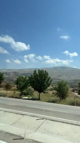 A roadside view shows a clear blue sky with scattered white clouds. A highway runs diagonally from the bottom left to the center right of the frame, bordered by a concrete barrier and a metal railing. Beyond the highway, a line of trees, some with green foliage and one with red leaves, are visible. In the background, rolling hills with sparse vegetation and a winding road are present. The overall environment appears to be a rural, mountainous landscape on a sunny day. The video appears to be filmed from a moving vehicle.