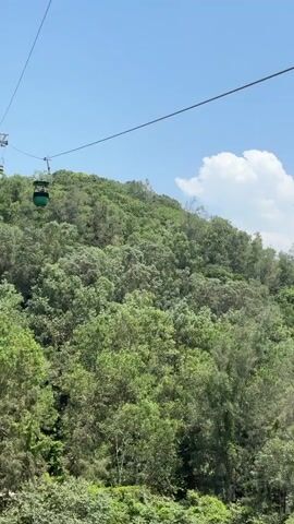 A green cable car travels along a cable over a lush, tree-covered mountainside under a blue sky with a few white clouds. The cable car is moving from left to right across the frame.