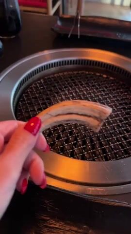 A person is placing a long, thin, beige-colored strip onto a circular metal grill. The grill is part of a built-in tabletop cooking setup with a stainless steel surround. The person's hand is visible, with red nail polish. The background shows a dark wooden tabletop with various items including a small black dish and tongs. The lighting is warm, suggesting an indoor restaurant setting.