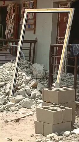 The scene depicts a partially demolished or deconstructed structure, possibly a building. Debris, including loose rocks and fragments, is scattered on the ground. A stack of concrete blocks is positioned in the foreground. Leaning against the debris is a rectangular frame, possibly a door or window frame, with a white or light-colored border and a black interior outline.

A man walks past the frame, glancing toward the camera as he passes. He is wearing a white t-shirt with a logo. Behind the debris and frame, there is a building with a visible shelf and what appear to be items for sale or display. Some metallic railings are also visible. The environment suggests ongoing construction, renovation, or demolition work.