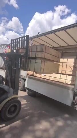 A yellow Komatsu forklift is positioned alongside a white truck trailer, its forks extended into the trailer. On the forks are several large, rectangular wooden crates stacked on top of each other. A person is visible operating the forklift, seated on the left side. The trailer is filled with more similar wooden crates. The scene takes place outdoors under a bright, partly cloudy sky, with trees and power lines visible in the background. The forklift appears to be in the process of loading or unloading the crates from the trailer.