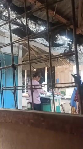 The video shows an outdoor scene through a barred window. In the background, a person wearing a pink shirt and a baseball cap is standing at a counter, seemingly preparing food. To the right of the frame, a woman wearing a patterned top and a dark headscarf is looking towards the person at the counter. Further to the right, a young boy in a red shirt is visible. The environment appears to be a makeshift outdoor area with a corrugated metal roof and netting overhead. The lighting suggests it is daytime.