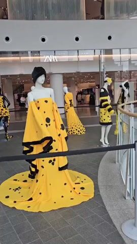 The video shows a display of mannequins dressed in yellow and black polka dot outfits inside a shopping mall. The mannequins are arranged in a semi-circle, with one large, elaborate gown in the foreground. Behind the mannequins, there is a Zara store visible. A banner in French, with the logo "ISA GOSSELIES", is displayed prominently in the center of the scene. The floor is tiled with a grey pattern. The overall lighting suggests an indoor, well-lit environment.