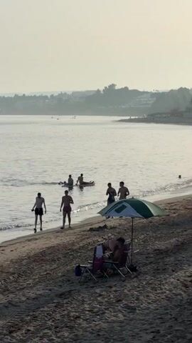 The video opens on a beach with calm ocean water. Several people are visible. In the foreground, two figures sit under a beach umbrella. Further down the beach, four young men are walking towards the water. In the ocean, two people are on a paddleboard. Another person is on a separate paddleboard further out. Additional people are visible on paddleboards or kayaks in the distance. The sky is hazy, and the background shows a coastline with vegetation and structures. The video pans slightly to the right, capturing more of the beach and the water.