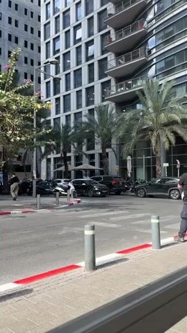 The video shows a street scene with modern buildings and parked cars. There are palm trees lining the street. A few cars are visible, some moving and some parked. A person is walking across the street. The sky is clear and the lighting suggests it is daytime. The overall environment appears to be an urban area with contemporary architecture.