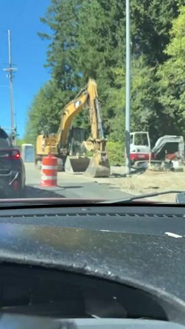 The video shows an excavator at a construction site on a sunny day. The excavator is yellow and has a large bucket. There are traffic barrels and construction equipment in the background. The video is shot from inside a vehicle, looking out at the construction site.