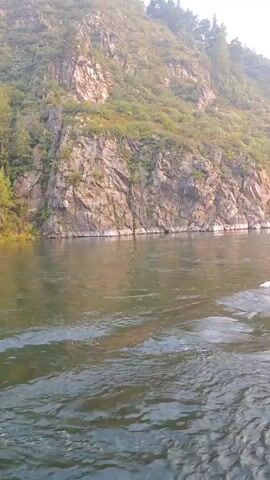 The video shows a boat moving through a river. The riverbanks are lined with steep, rocky cliffs covered in green vegetation. The water is choppy, with visible wakes from the boat. The sky is hazy and appears to be early morning or late afternoon.