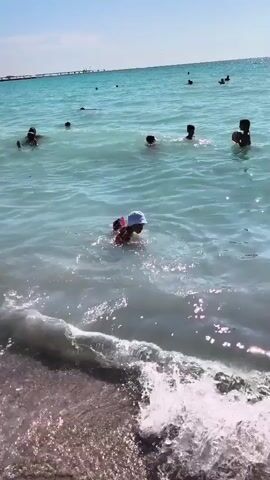 A group of people are in the ocean on a sunny day. The water is clear and blue. In the foreground, a small child wearing arm floaties and a hat is splashing in the water. Further out, several other individuals, including children and adults, are swimming and playing. The waves are gently lapping at the shore. The sky is clear and blue, and the sun is shining brightly.