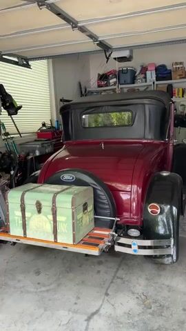 The video shows the back and side of a vintage maroon Ford automobile parked in a garage. The car has a black soft top and black fenders. A trunk is strapped to the back of the car. The camera moves from the back to the side of the car, where a hand opens the driver's side door. The interior of the car is visible through the open door. Various items are stored on shelves and in containers around the garage. The floor of the garage is concrete.