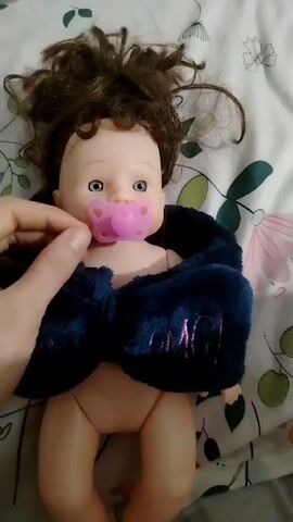 The video shows a doll lying on a patterned blanket. The doll has brown, messy hair, blue eyes, and a painted-on mouth with lipstick. It is partially covered by a dark blue fuzzy wrap with the word "MAGIC" embroidered on it. A hand enters the frame and removes a pink pacifier from the doll's mouth. The doll then makes a crying sound. The hand then holds the pacifier in front of the doll's mouth again. The blanket has a floral pattern.
