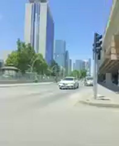 The video shows a street scene with several cars driving down the road. On the left side of the road, there are tall buildings and trees. On the right side of the road, there is an elevated structure, possibly a bridge or overpass. The sky is clear and blue, indicating it is daytime. The video appears to be filmed from a moving vehicle. There are no discernible interactions or specific actions taking place that would require further detail.