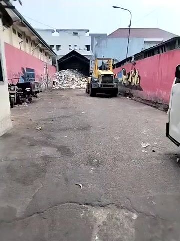 The video shows a loading dock area. In the foreground, a white truck is parked on the right, partially obscuring the view. A forklift is operating in the background, near a large pile of debris. The area appears to be industrial or a waste disposal site, with buildings visible on either side of the loading dock. The ground is paved, and there is scattered debris. The video is shot from a moving perspective, suggesting it was recorded by someone walking or driving through the area. There are no people clearly visible interacting with the forklift or the debris pile. The overall impression is of a functional, albeit somewhat unkempt, industrial environment.