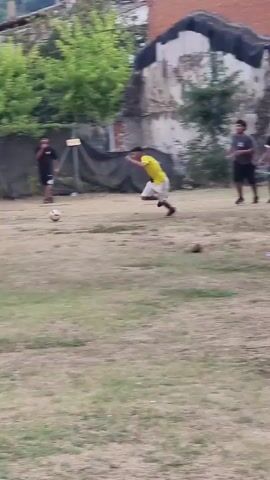The video shows a group of boys playing soccer in an outdoor area. The ground is covered with dry grass and some patches of dirt. In the background, there are buildings, trees, and a fence. The boys are wearing casual clothing, and some of them are wearing soccer jerseys. They are actively engaged in playing the game, with one boy dribbling the ball and others attempting to intercept or defend. The camera is positioned at a distance, providing a wide view of the scene. The lighting suggests it is daytime, possibly late afternoon due to the shadows.
