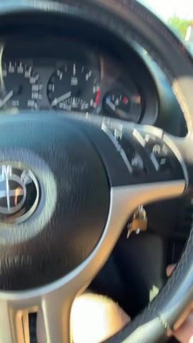 A hand is seen turning the ignition of a BMW. The dashboard lights up, showing various warning lights and the odometer reading 16650. The car then starts, and the speedometer needle rises slightly. The video is filmed from the driver's perspective, looking at the steering wheel and dashboard. Outside, a street with houses and trees is visible.