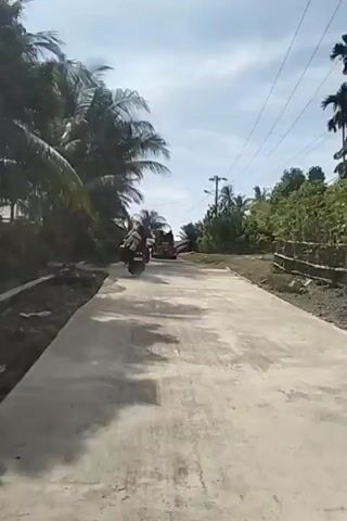 A drone shot follows a paved road in a rural area with palm trees and vegetation on either side. A motorcycle carrying two passengers drives ahead on the road, followed by a red three-wheeled vehicle also carrying passengers. The vehicle appears to be a motorized tricycle with a covered passenger area. The sky is partly cloudy.