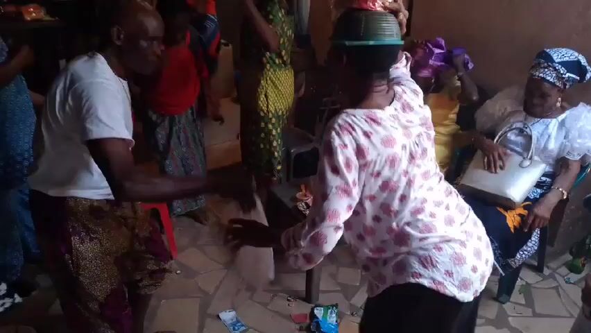 Inside a room with painted walls, a group of people are gathered, seemingly celebrating. A woman in a pink and white floral top is balancing a decorative object on her head. Around her, others are dancing and clapping. A man in a white t-shirt and patterned skirt is particularly animated, waving a white cloth. An older woman with a head wrap is seated in a chair, holding a purse. A table nearby holds items like bottles and cash. The room has a casual, intimate atmosphere.