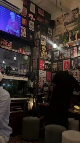 The video pans across a dimly lit room decorated with numerous posters and framed pictures covering the walls. A person in a black shirt stands facing away from the camera, partially obscuring another person seated at a small table. In the foreground, there are several round, cushioned stools. The environment appears to be a bar or cafe, with shelves filled with bottles visible in the background.