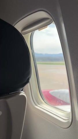 The video shows a view from an airplane window. The wing of the plane, painted with red and white stripes, is partially visible below the window. Outside, a runway stretches into the distance. Beyond the runway, there's a green patch of land, followed by a town or a settlement. In the far background, snow-capped mountains can be seen under a cloudy sky. The interior of the plane is visible, including the window frame and a portion of a seat with a black headrest.
