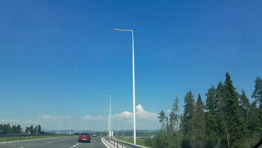 The video shows a view from a vehicle traveling on a highway. The sky is clear and blue with a few scattered white clouds. Tall white lamp posts are visible along the side of the road, and a line of trees borders the highway on the right. Several cars and a bus are seen on the highway. The vehicle appears to be moving at a moderate speed.