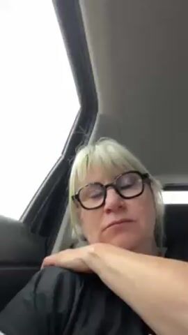 A woman with short blonde hair and glasses is asleep in a car. Her eyes are closed, and she is leaning against the side of the car. The interior of the car is visible, including the roof and the black seat. The car is in motion, with background music playing. The scene is brightly lit from outside the car.