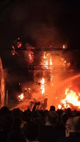 In the video, a large crowd watches as several intricate wooden structures are engulfed in flames at night. The flames are bright orange, creating a hazy atmosphere filled with smoke. The structures appear to be interconnected by scaffolding and are adorned with additional small light elements. The crowd, seen mostly from the back, consists of various individuals, some holding up phones to capture the spectacle. The background is dark, with faint outlines of buildings visible on the left side of the frame. A few dangling wires are barely discernible against the night sky.